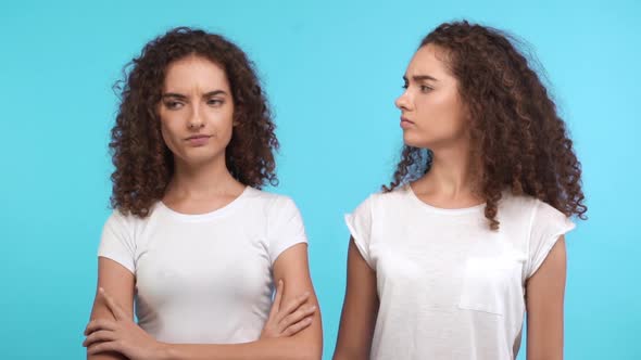 One Dissapointed Female Curly Caucasian Twin Standing on Blue Background While Another Smiling