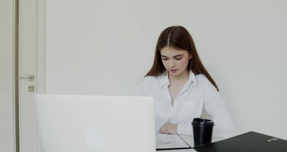 Cheerful Girl Talking Online on Laptop and Noting at Workplace in Light Office