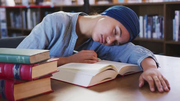 Asian female student wearing a blue hijab sitting at a desk with an open book and sleeping