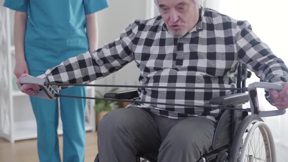 Elderly Grey-haired Caucasian Man in Wheelchair Exercising As Professional Unrecognizable Nurse