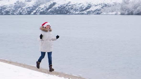 A Beautiful Woman Runs Along the River in Winter Santa Claus' Christmas Hat on Her Head