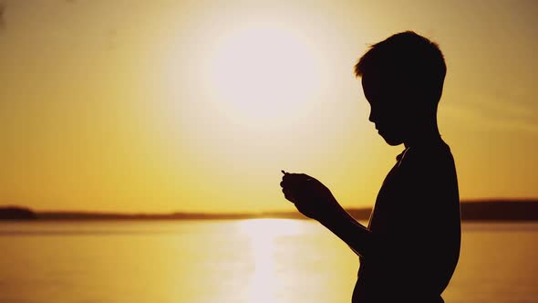 Silhouette of a child by the river. Paper boat is in the hands of the child.