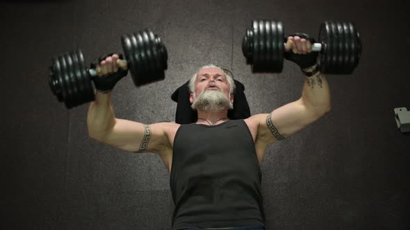 An overhead view of an athletic gray-haired man with a beard and tattoos, training his chest muscles