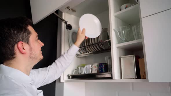 Young adult male Caucasian ethnicity 30s open a cupboard with clean dishes and puts plates in it