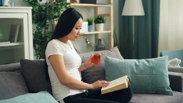 Good-looking Asian Woman Is Reading Book Enjoying Modern Literature Sitting on Sofa at Home Drinking