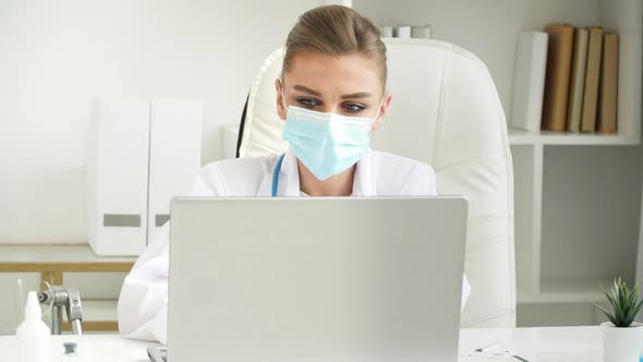 Female Doctor in Mask Works at Laptop in Clinic