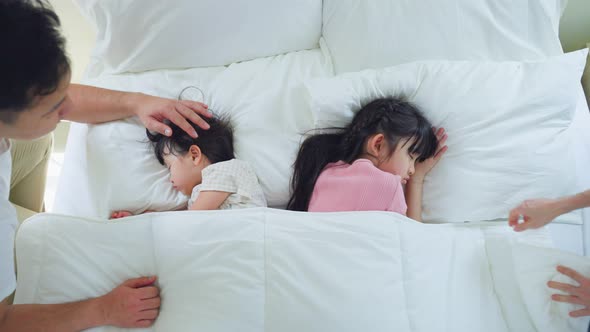 Asian mother and father puts blanket on comfortable asleep napping little baby kid on bed.