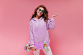 Charming girl in purple hoodie and white pants smiles and holds colorful longboard. Happy woman in - PhotoDune Item for Sale