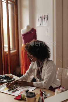 Stylish curly brunette dark-skinned lady in white blouse smiles sincerely and paints stylish clothe / Photodune, Misc