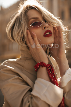 Beautiful sexy self-confident blonde woman in beige trench coat, red sunglasses looks into camera a / Photodune, Misc