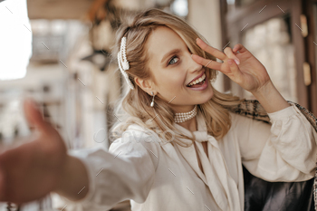 Cool beautiful blonde woman in white stylish blouse and pearl jewelry smiles, shows peace sign and / Photodune, Misc