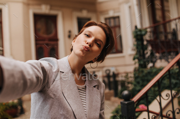 Brown eyed lady in grey jacket takes selfie outside and blows kiss. Charming woman in suit and tee / Photodune, Misc
