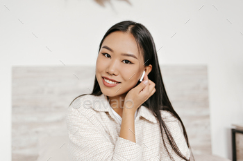 Brown-eyed brunette Asian woman in good mood listens to music in wireless headphones and smiles sin / Photodune, Misc
