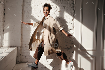 Amazed black young woman in trench coat looking at camera. Full length view of african american mod / Photodune, Misc