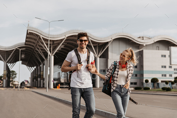 Brunette man in white tee and girl in plaid shirt smiles and walks with backpacks along road. Trave / Photodune, Misc