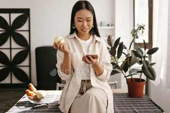 Attractive Asian woman in beige cardigan, stylish pants and white t-shirt sits on table on kitchen, / Photodune, Misc