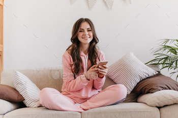 Photo made in homely atmosphere. Cute brunette in pink pajamas uses her phone and smiles sweetly / Photodune, Misc