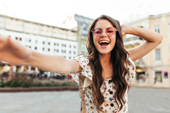 Joyful brunette curly girl in white floral top takes selfie outside. Happy young woman in pink sung / Photodune, Misc