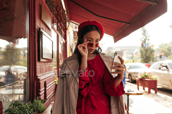 Attractive brunette Asian woman in stylish beret, red dress and beige trench coat puts off sunglass / Photodune, Misc