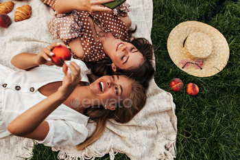 Joyful brunette and happy blonde women in summer stylish outfits hold peach and have picnic outdoor / Photodune, Misc