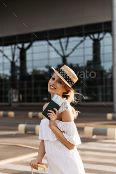 Pretty blonde woman in hat smiles sincerely. Girl in white dress poses near airport. Tourist holds / Photodune, Misc