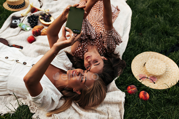 Attractive tanned girls in summer outfits lay on linen rug and have picnic outdoors. Young women in / Photodune, Misc
