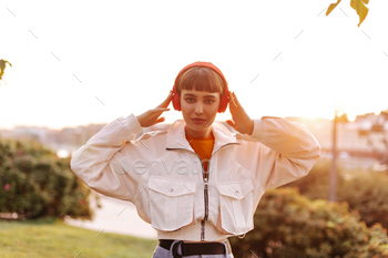 Attractive girl in white stylish jacket listens to music in red headphones. Short-haired woman look / Photodune, Misc