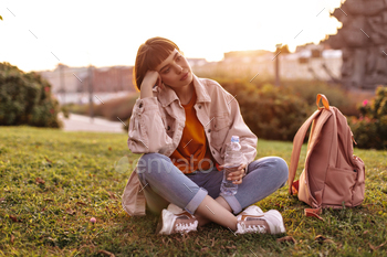 Sad young woman sits on grass in city during sunset. Short-haired girl in jeans and pink denim jack / Photodune, Misc