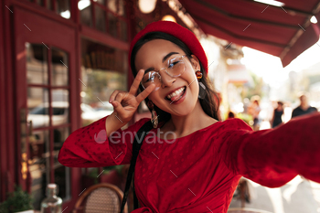 Joyful optimistic woman in red dress, bright beret and eyeglasses takes selfie, shows peace sign an / Photodune, Misc