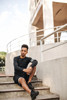 Curly bearded brunet dark-skinned man in shorts and long-sleeved t-shirt looks into camera and sits / Photodune, Misc