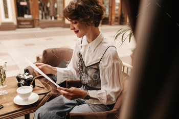 Short-haired girl with bright lips smiles and sits in cafe. Young lady in modern shirt and jeans re / Photodune, Misc