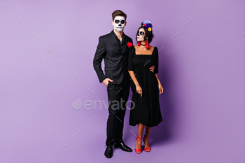 Full-length photo of guy and girl in elegant black outfit and Halloween masks posing on purple back / Photodune, Misc