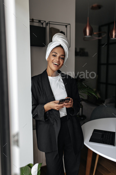 Excited lady in black suit smiles and leans on wall in office. Dark-skinned beautiful woman in styl / Photodune, Misc