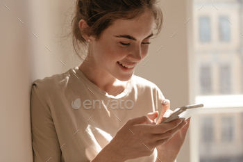 Close-up portrait of young girl looking at phone in light room. Cute brunette with beige t-shirt, h / Photodune, Misc