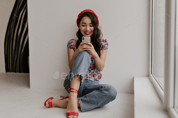 Cool brunette curly Asian woman in red beret, floral blouse and denim pants leans on white wall, si / Photodune, Misc