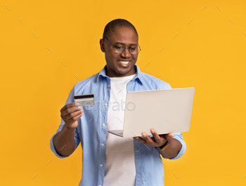 ing. Happy millennial muscular handsome african american male in glasses holding laptop and credit card, isolated on yellow background, studio shot