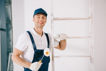 handsome young painter in uniform smiling at camera while standing near ladder and holding paint / Photodune, Misc