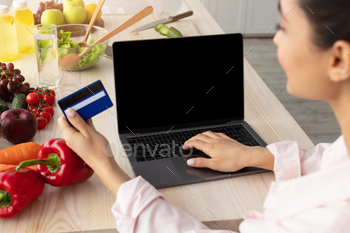 nizable positive woman using laptop pc with empty black screen for mock up, sitting at kitchen counter, holding and showing credit card, paying online