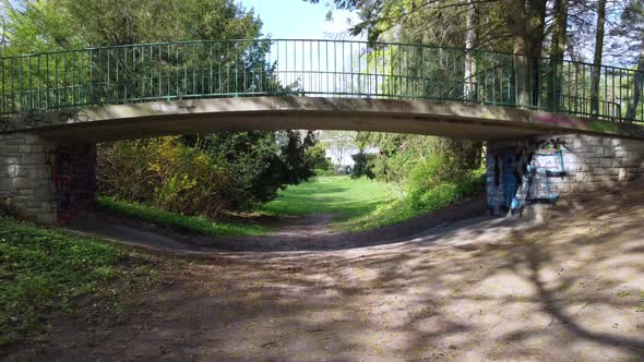 Along a path under a footbridge through into a park. Unbelievable aerial view flight fly forwards dr