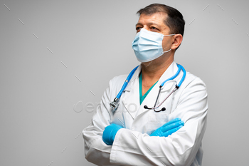 Senior male doctor in face mask standing against grey background / Photodune, Misc