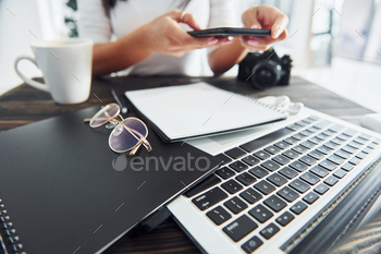 Holding phone. Young female freelancer working indoors in the office at daytime. Laptop and notepad / Photodune, Misc