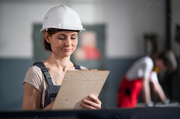 Portrait of woman worker with helmet and clipboard indoors in factory making notes / Photodune, Misc