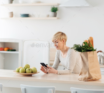  modern devices and app. Good offer and add, social networks. Smiling blonde mature woman typing message in tphone on kitchen interior, empty space