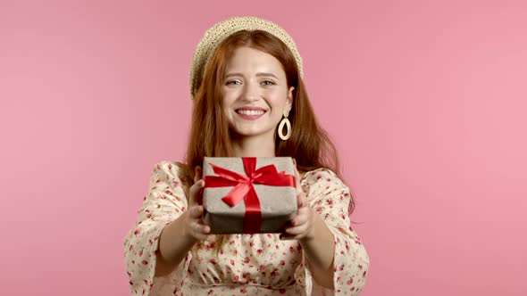 Cute Young Woman in Dress Holding Gift Box and Gives It By Hands To Camera on Pink Wall Background