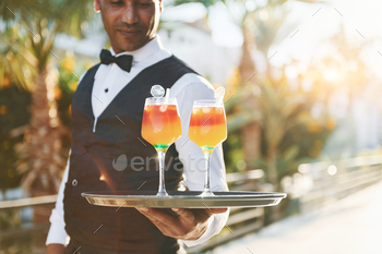 Holds cocktails. Black waiter in formal clothes is at his work outdoors at sunny daytime / Photodune, Misc