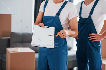 Holds notepad with document. Two young movers in blue uniform working indoors in the room / Photodune, Misc