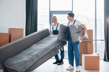 Carrying heavy sofa. Two young movers in blue uniform working indoors in the room / Photodune, Misc