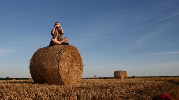 blonde girl is sitting on rolled haystack with cup of coffee in field in sunset time