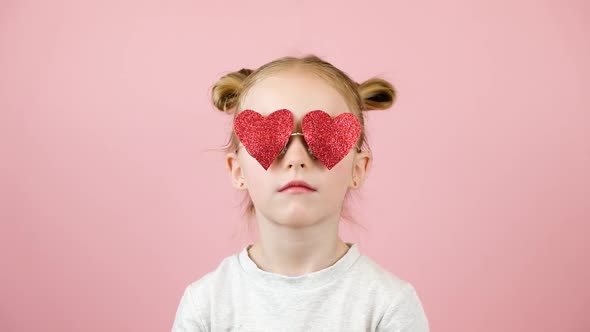 Funny Little Blonde Girl Smiling and Playing with Red Heart Shape Sunglasses on Pink Background