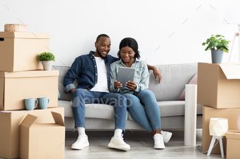 hile Sitting On Couch After Moving To New Home, Cheerful African American Spouses Shopping Online On Tab Computer Or Browsing Internet, Copy Space